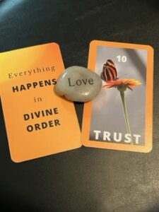 Two gold cards lie flat with a rock between them engraved with the word love. The card on the left has the text Everything happens in divine order. The card on the right has the number 10 at the top in white letters above an image of a orange and black butterfly on top of a gold flower. at the bottom the word trust is in white letters.