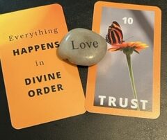 Two gold cards lie flat with a rock between them engraved with the word love. The card on the left has the text Everything happens in divine order. The card on the right has the number 10 at the top in white letters above an image of a orange and black butterfly on top of a gold flower. at the bottom the word trust is in white letters.