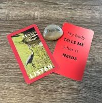 Two red cards, 3x5 in. Card on the left with a great blue heron and the word Listen in white letters. A rock with thank you engraved sits between the two cards. The card on the right read - My body tells me what it needs
