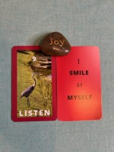 Two red 3 x 5 cards next to each other with a rock with the word Joy engraved sits in the middle. On the left card an image of a great blue heron near a lake is between the #9 and the word listen, both in white. Text on the other card reads I smile at myself.