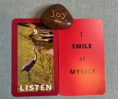 Two red 3 x 5 cards next to each other with a rock with the word Joy engraved sits in the middle. On the left card an image of a great blue heron near a lake is between the #9 and the word listen, both in white. Text on the other card reads I smile at myself.