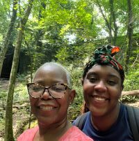 Two brown skinned women wearing glasses stand with a forest of trees behind them.