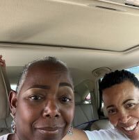 2 brown skinned women with freshly made up faces are seated in a car. Barbara on the left has one eyebrow raised with no smile. Regenna on the right smiles.