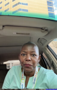 Middle-aged pecan brown woman with closely cropped hair sits inside a car with beige interior.