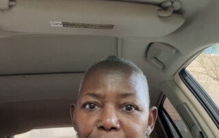 Middle-aged pecan brown woman with closely cropped hair sits inside a car with beige interior.