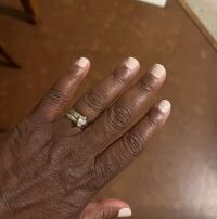 A pecan brown colored left hand against the same colored background shows light pink nail polish, an wedding ring set is on the fourth finger.