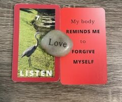 Two red 3 x 5 cards next to each other with a rock with the word Joy engraved sits in the middle. On the left card an image of a great blue heron near a lake is between the #7 and the word listen, both in white. Text on the other card reads My body reminds me to forgive myself in black text